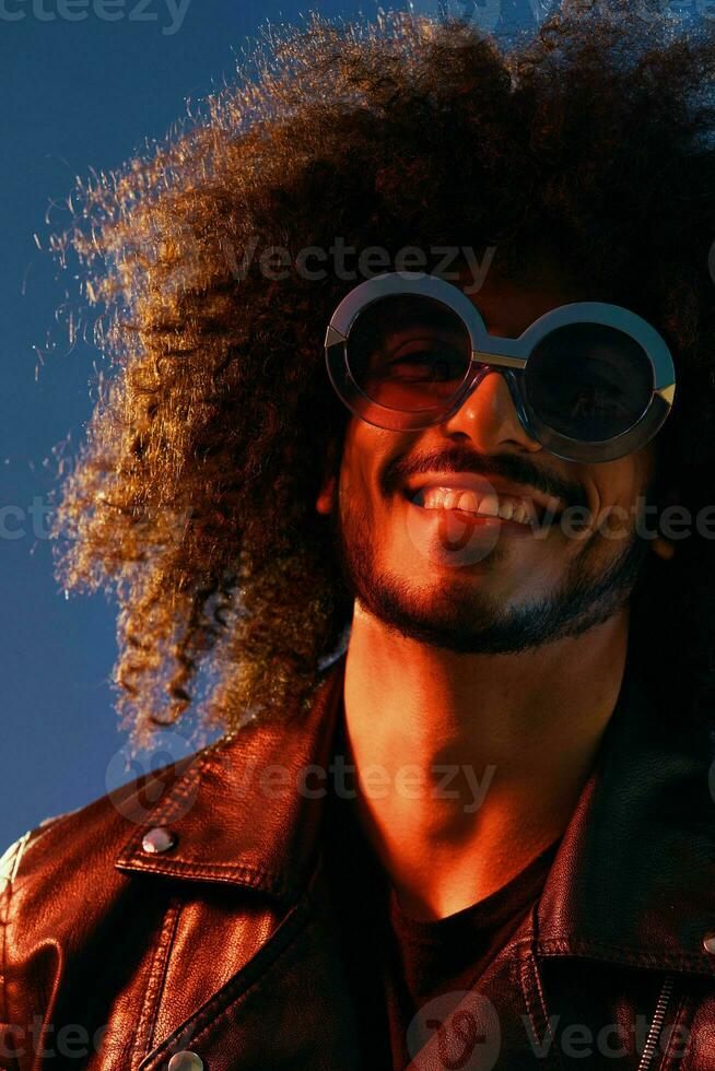 Portrait of a stylish man with curly hair with glasses smile on blue background multinational, color light, black leather jacket trend, modern concept. photo