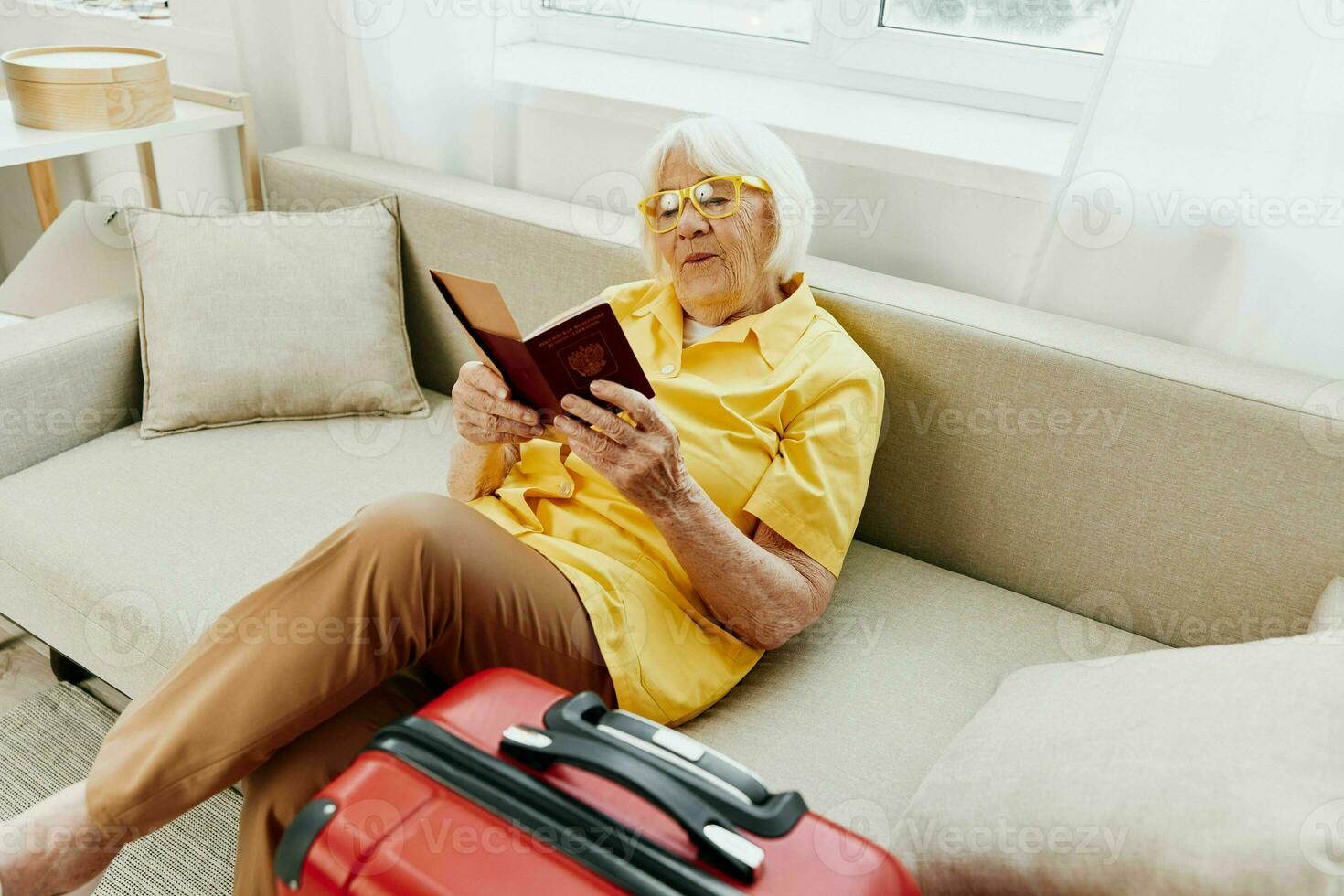 Happy senior woman with passport and travel ticket packed a red suitcase, vacation and health care. Smiling old woman joyfully sitting on the sofa before the trip raised her hands up in joy. photo