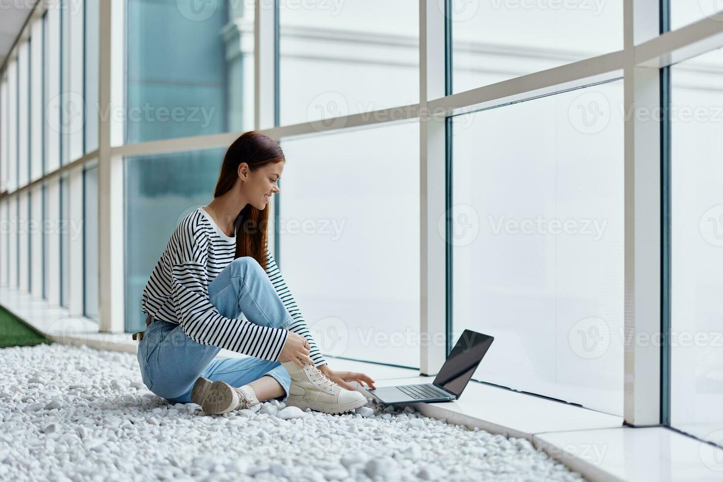 A woman smiling with teeth sits with a laptop by the big windows of a building in the city, the concept of work and leisure in the big city photo