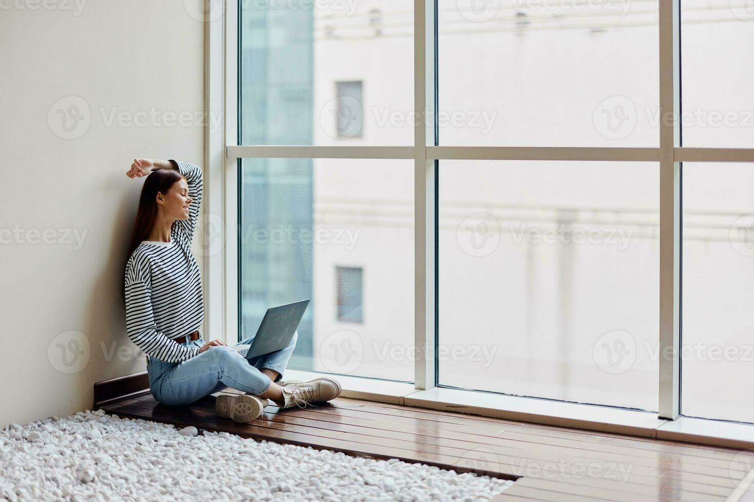A young woman sits on the floor with her laptop by the window working and studying in the big city photo