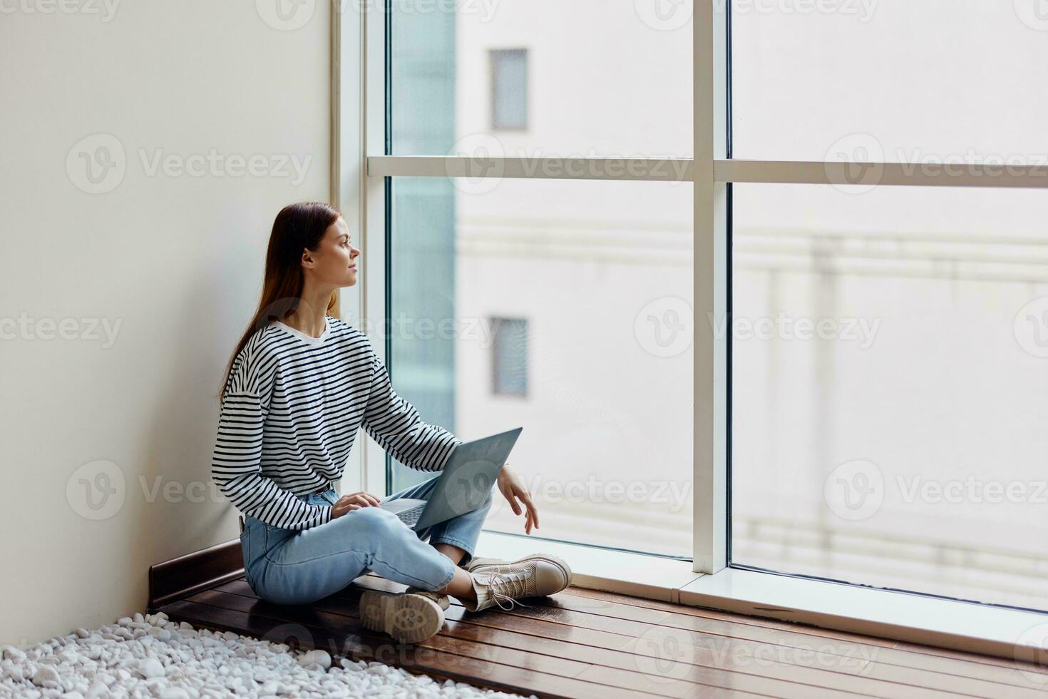 A woman freelancer sits with her laptop full-length at a large window, looking out at tall buildings in the city, work, training and shopping online photo
