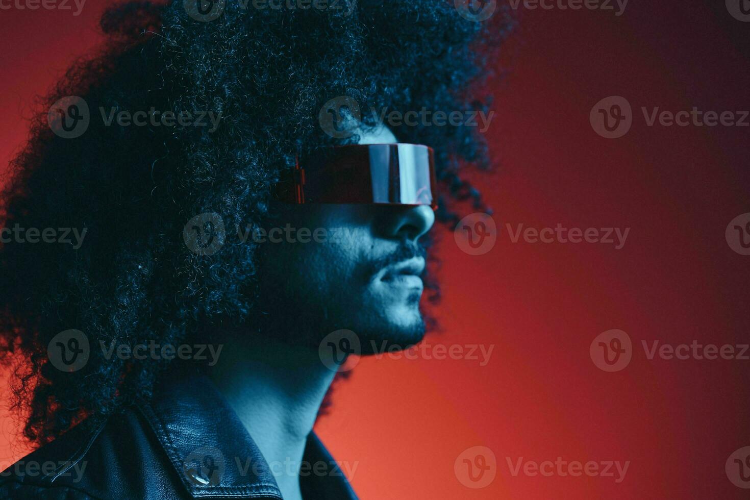 Portrait of fashion man with curly hair on red background with stylish glasses, multicultural, colored light, black leather jacket trend, modern concept. photo