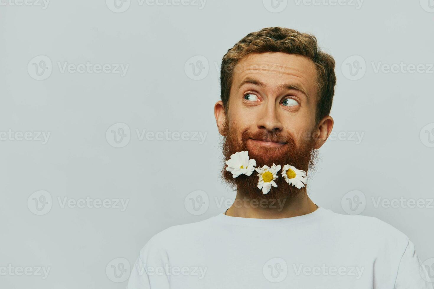 Portrait of a funny man in a white T-shirt with flowers daisies in his beard on a white isolated background, copy place. Holiday concept and congratulations. photo