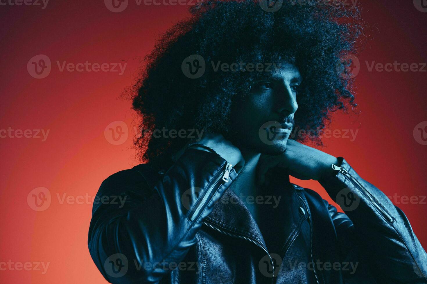 Portrait of fashion man with curly hair on red background multinational, colored light, black leather jacket trend, modern concept. photo