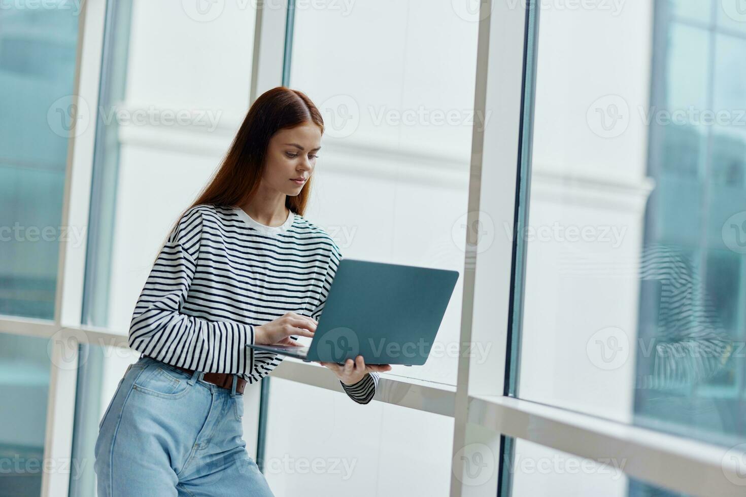 A woman standing near a large window in the city and holding a laptop in her hand looking out the window at the cityscape, a copy space photo