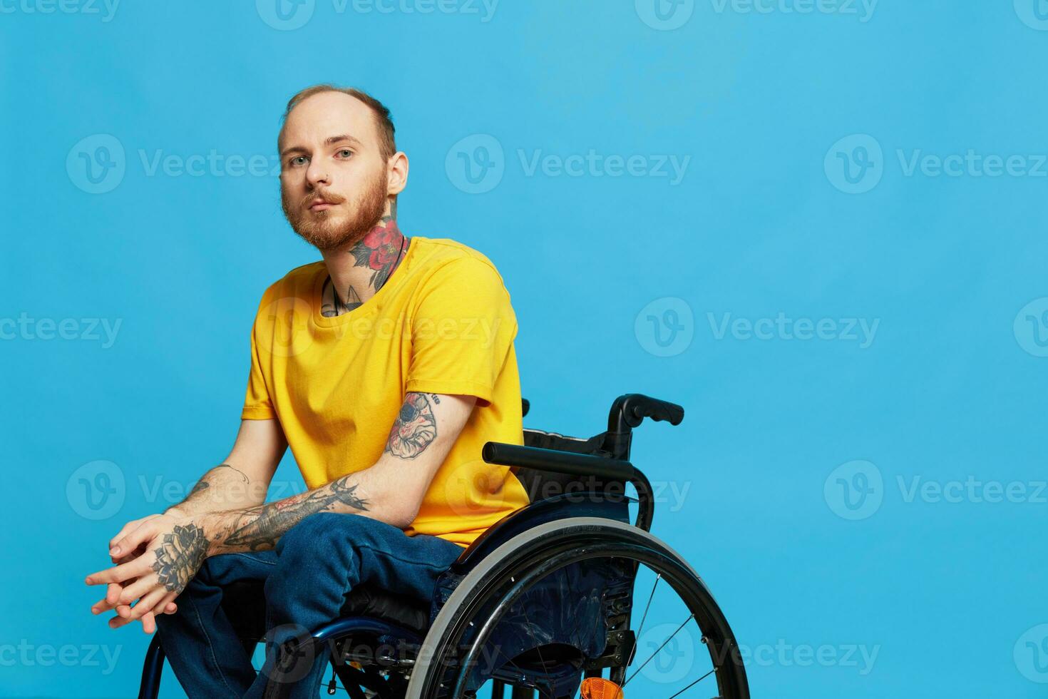 A man in a wheelchair problems with the musculoskeletal system looks at the camera in a t-shirt with tattoos on his arms sits on a blue studio background, full life, real person, health concept photo