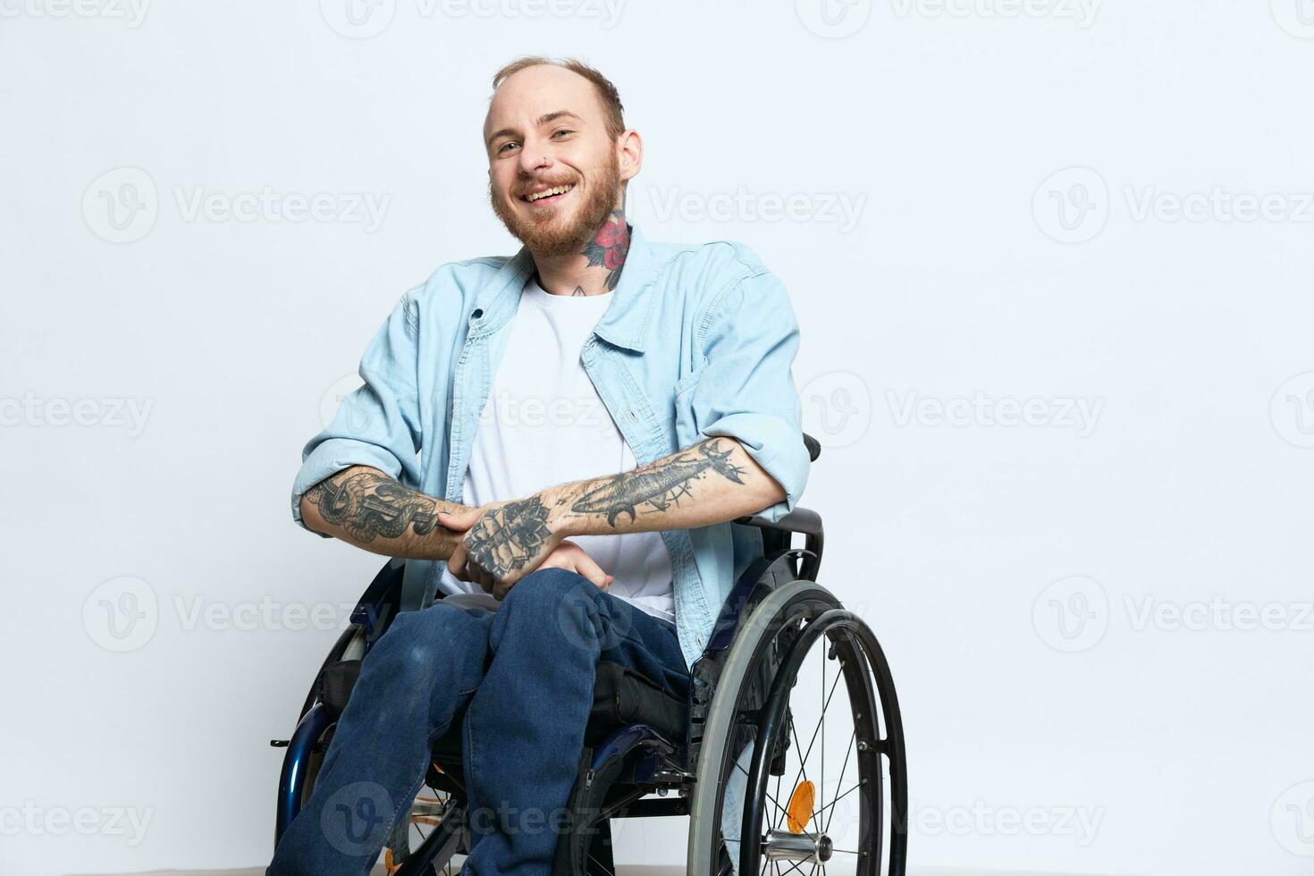 A man in a wheelchair and pointing a finger at, copy space, with tattoos on his hands sits on a gray studio background, health concept a person with disabilities, a real person photo