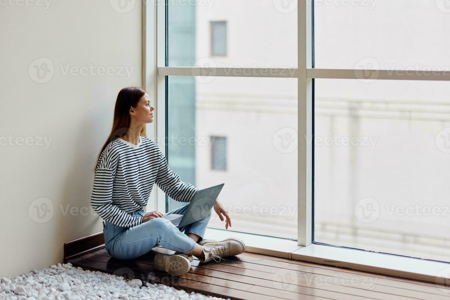 A woman freelancer sits with her laptop full-length at a large window, looking out at tall buildings in the city, work, training and shopping online photo