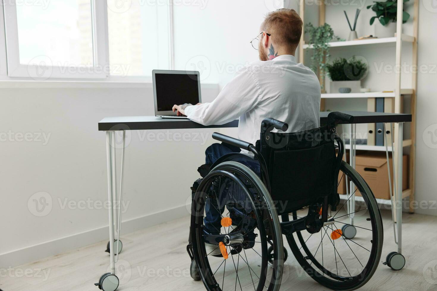 un hombre silla de ruedas empresario con tatuajes en el oficina trabajos a un ordenador portátil en línea, negocio proceso, un silla de ruedas de cerca, integración dentro sociedad, el concepto de trabajando un persona con discapacidades foto