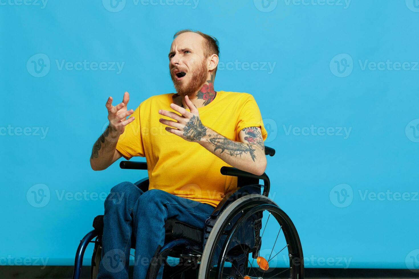 A man in a wheelchair in a T-shirt with tattoos on his arms sits on a blue studio background, a full life, a real person photo