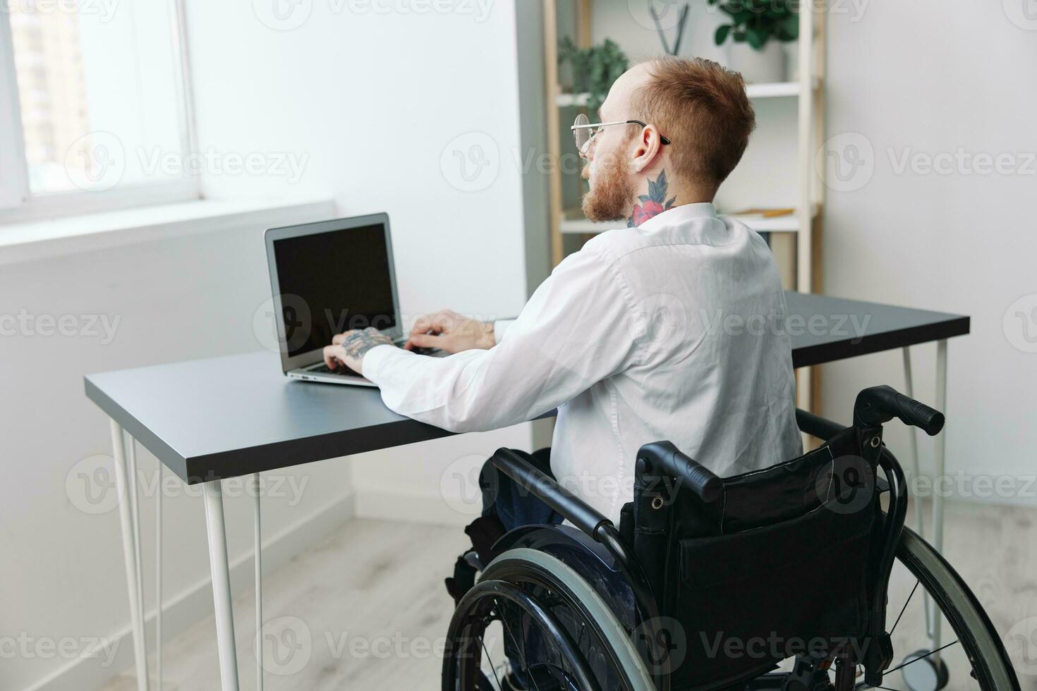 un hombre silla de ruedas empresario con tatuajes oficina trabajos a un ordenador portátil en línea, social redes y puesta en marcha, integración dentro sociedad, el concepto de trabajando un persona con discapacidades, ver desde el espalda foto