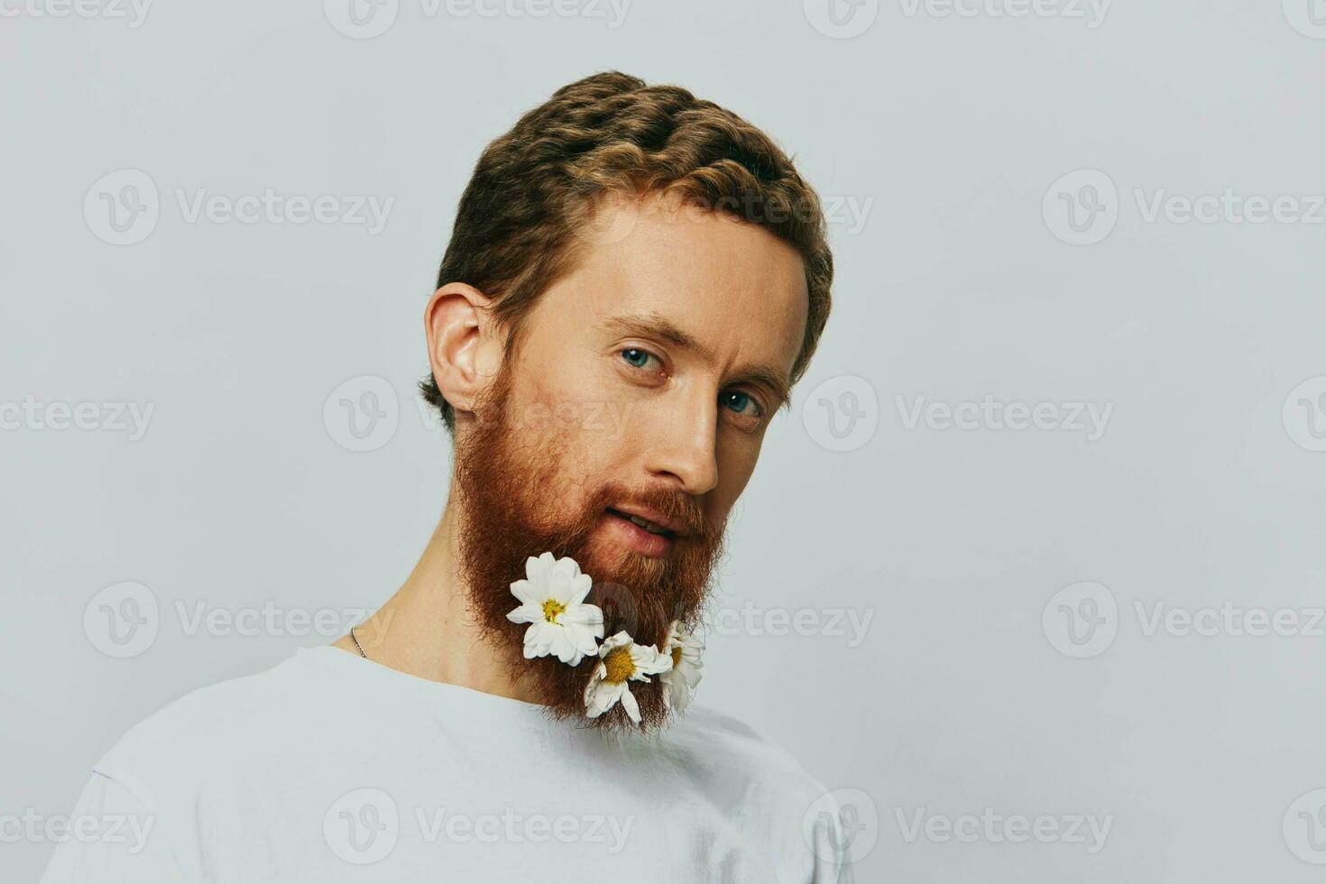 Portrait of a funny man in a white T-shirt with flowers daisies in his beard on a white isolated background, copy place. Holiday concept and congratulations. photo