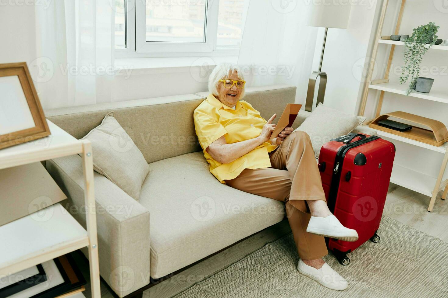 Happy senior woman with passport and travel ticket packed a red suitcase, vacation and health care. Smiling old woman joyfully sitting on the sofa before the trip raised her hands up in joy. photo