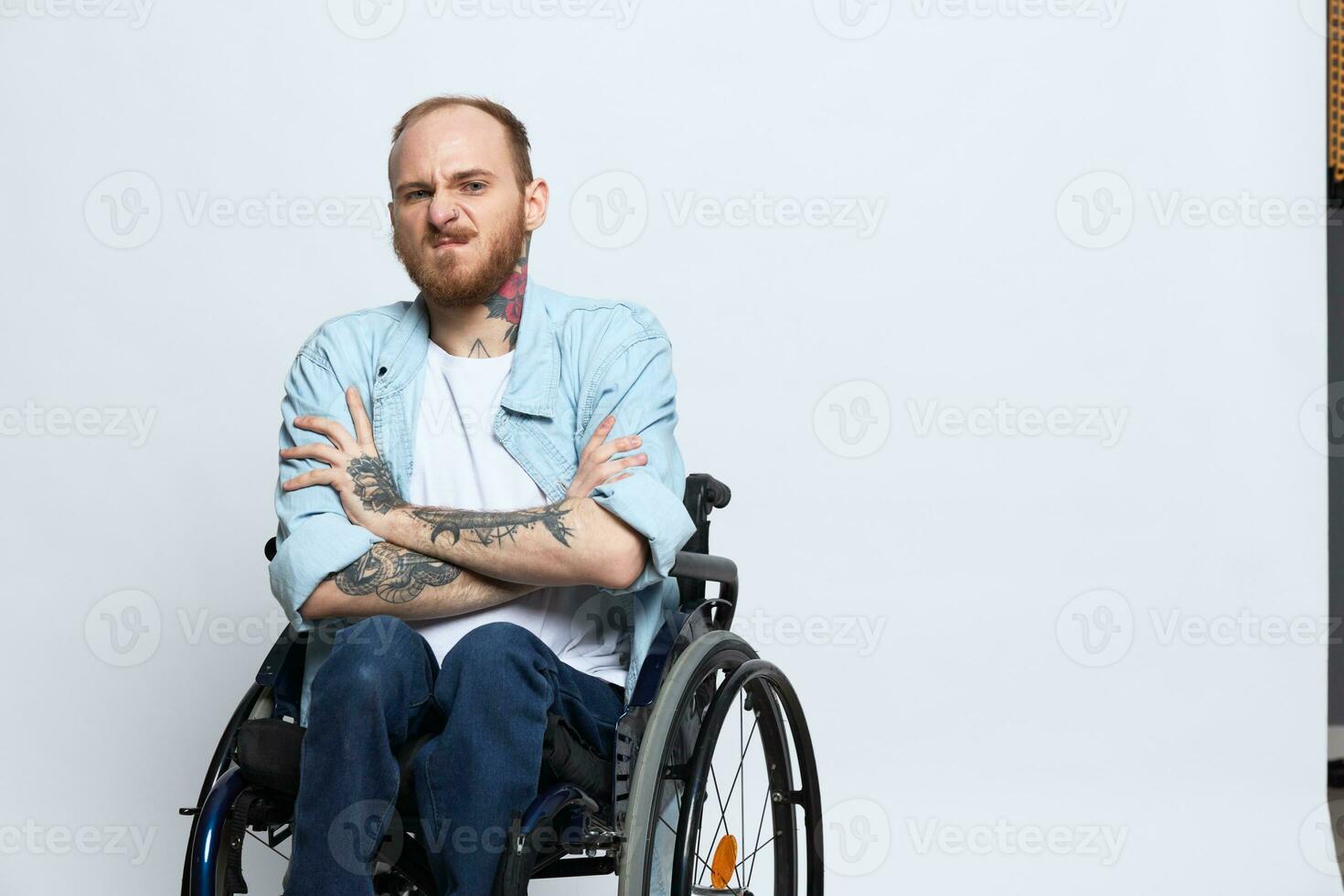 A man in a wheelchair looks at the camera anger and aggression, with tattoos on his arms sits on a gray studio background, health concept man with disabilities photo