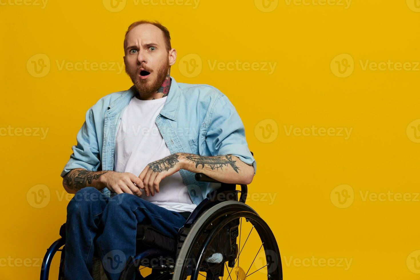 A man in a wheelchair surprised, with tattoos on his arms sits on a yellow studio background, the concept of health is a person with disabilities photo