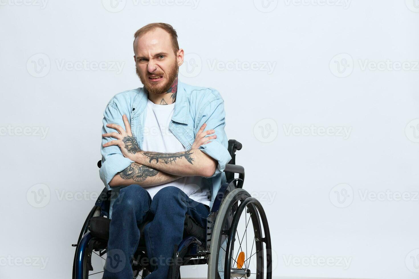 A man in a wheelchair looks at the camera anger and aggression, with tattoos on his arms sits on a gray studio background, health concept man with disabilities photo