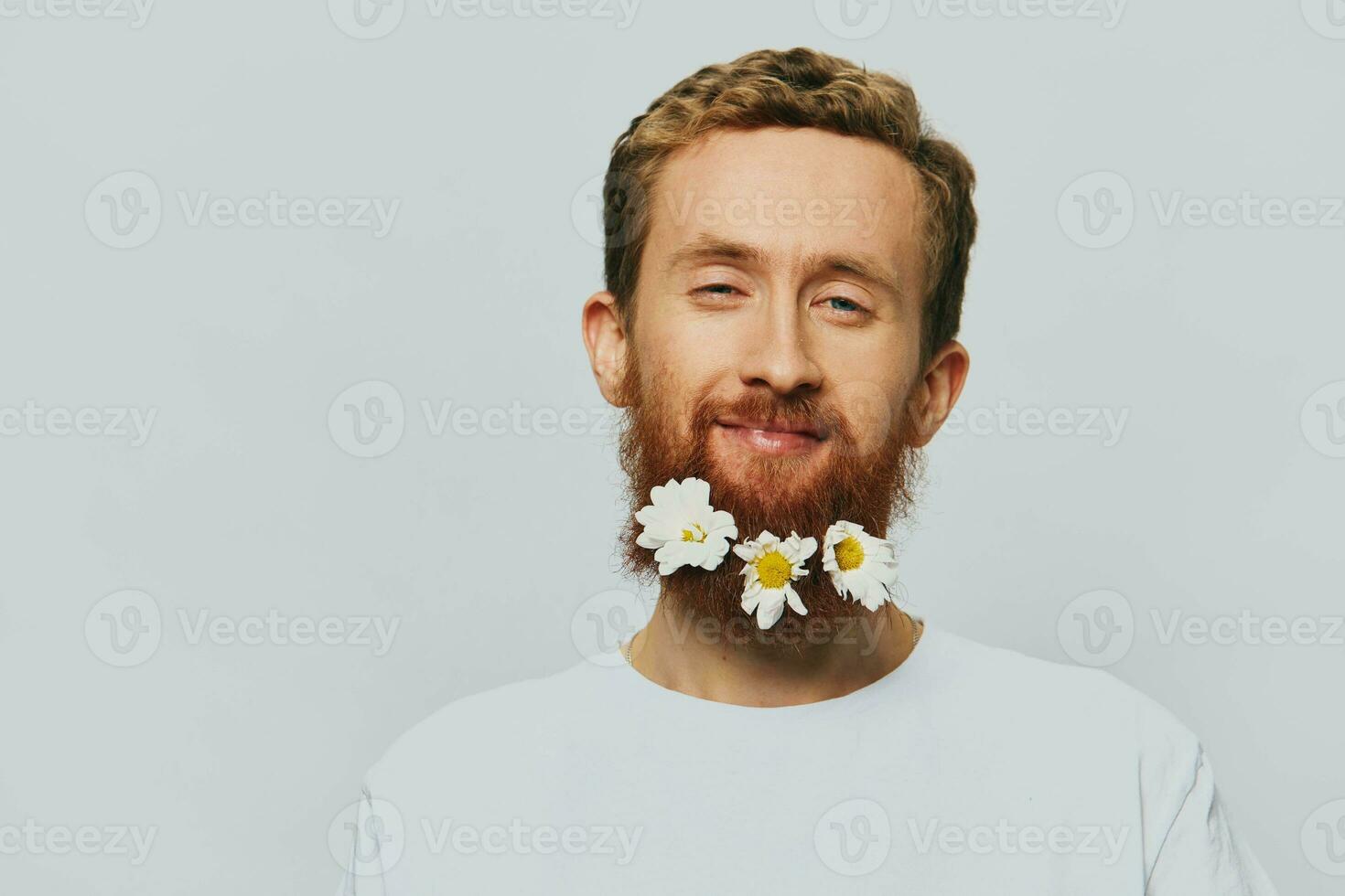 Portrait of a funny man in a white T-shirt with flowers daisies in his beard on a white isolated background, copy place. Holiday concept and congratulations. photo