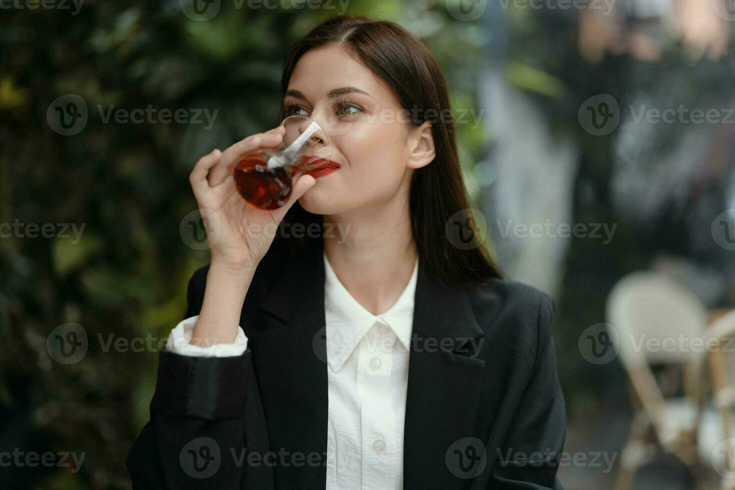 Business brunette woman with red lips smile with teeth drinks tea in a cafe from a Turkish glass mug in a white shirt and black fashion jacket on the street, summer travel, vacation in the city photo