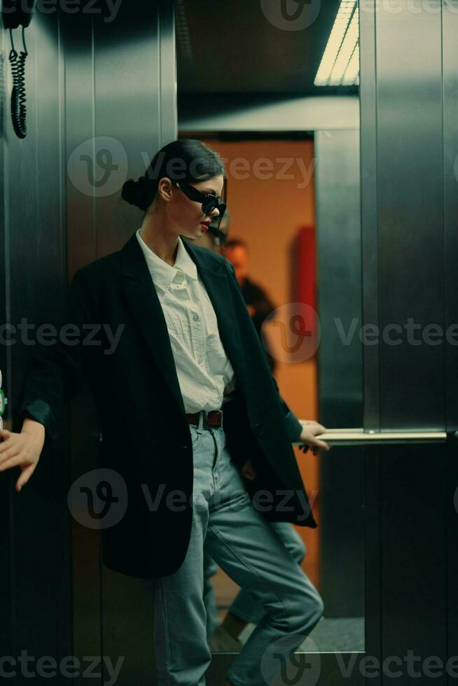 Stylish woman in black jacket and sunglasses posing in elevator, fashion model, dark cinematic light and color photo