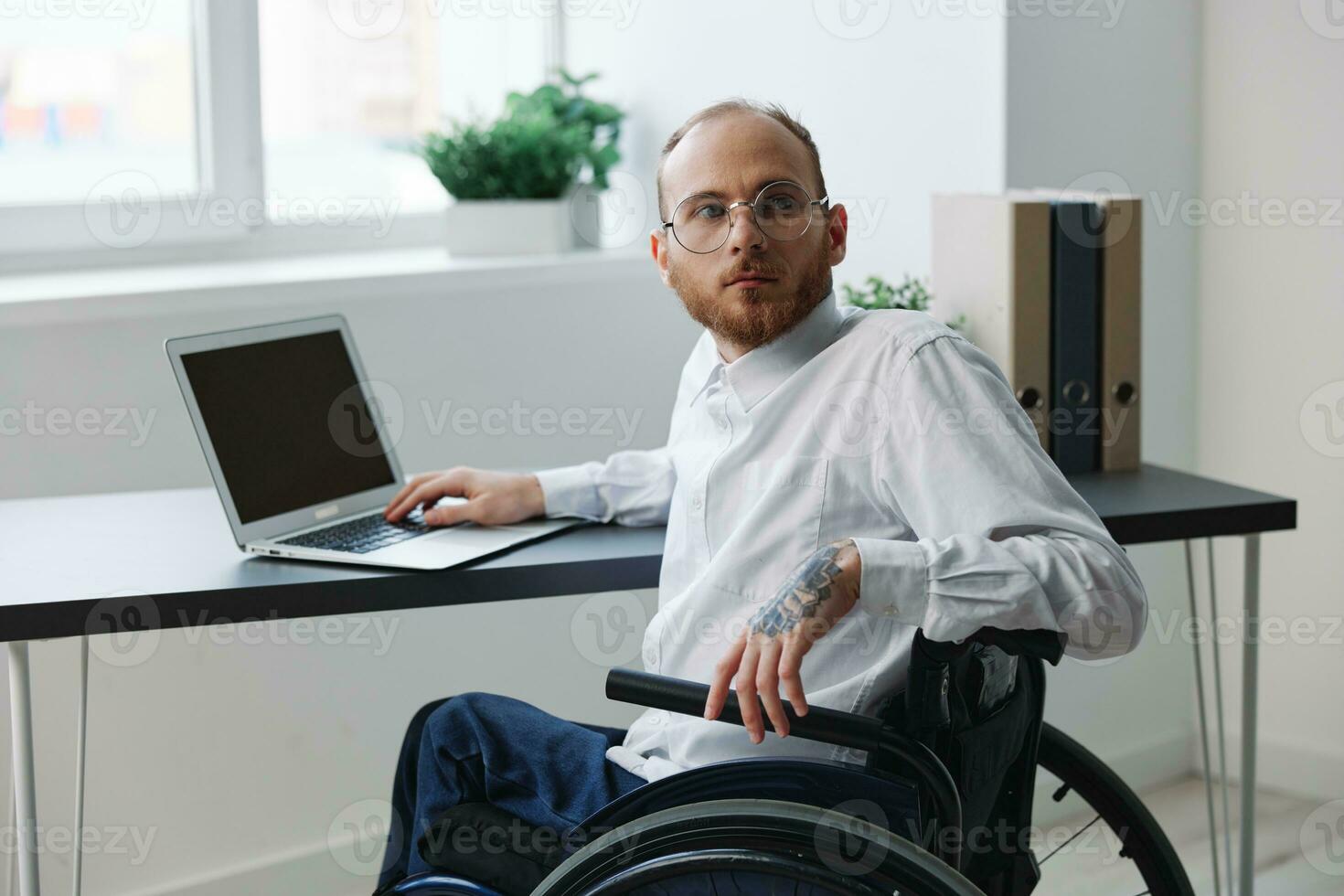 un hombre en un silla de ruedas mirando a el cámara empresario en el oficina trabajando en un ordenador portátil en línea, social redes y puesta en marcha, integración dentro sociedad, trabajo concepto hombre con discapacidades foto