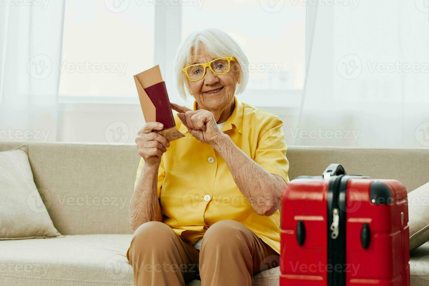 Happy senior woman with passport and travel ticket packed a red suitcase, vacation and health care. Smiling old woman joyfully sitting on the sofa before the trip raised her hands up in joy. photo