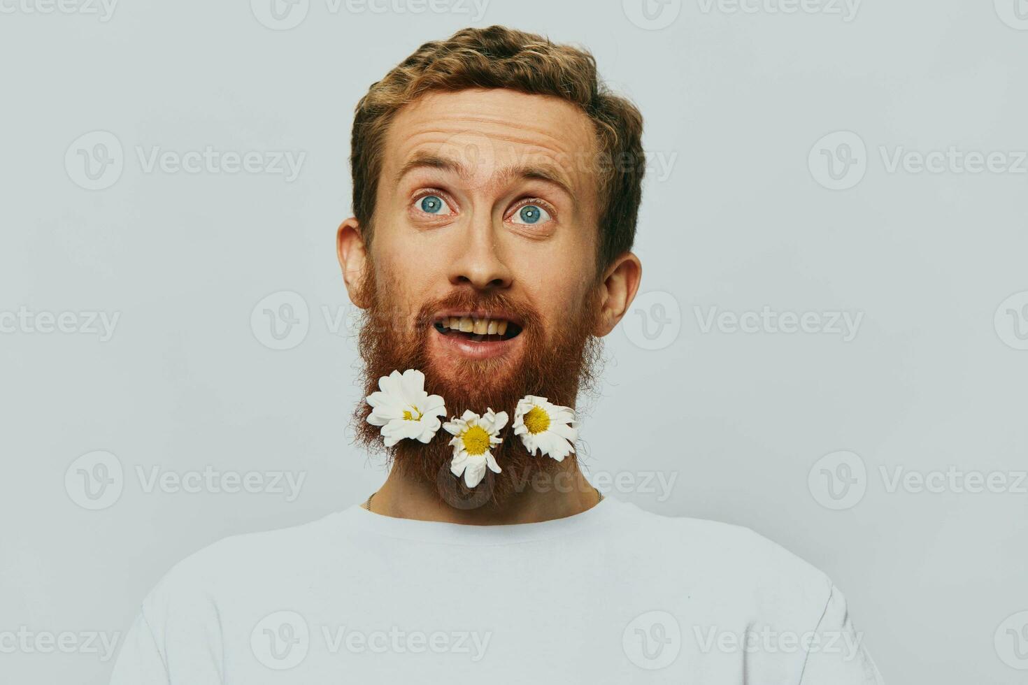 Portrait of a funny man in a white T-shirt with flowers daisies in his beard on a white isolated background, copy place. Holiday concept and congratulations. photo