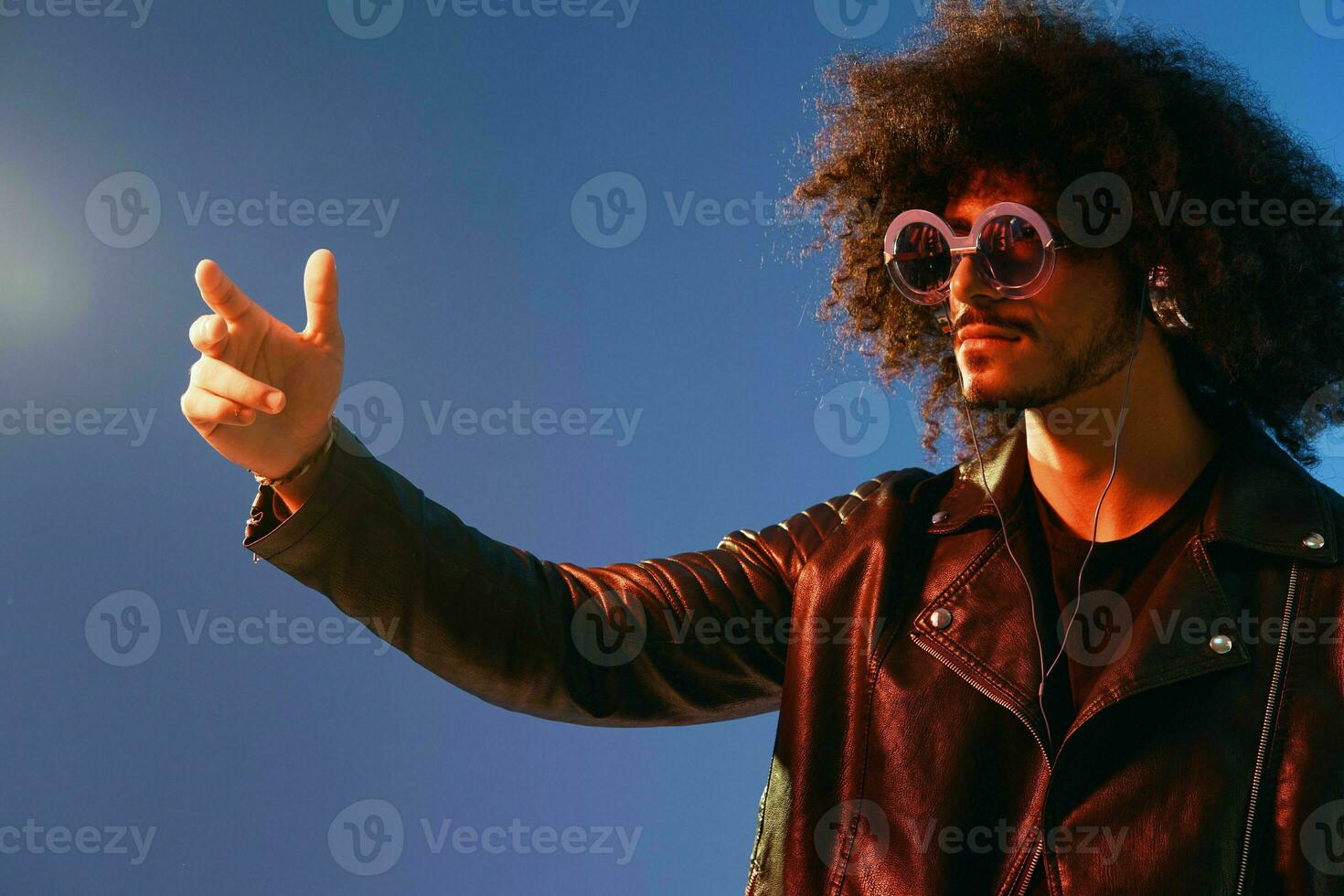 Portrait of a stylish man with curly hair with glasses and headphones on a blue background multinational, colored light, black leather jacket trend, modern concept. photo