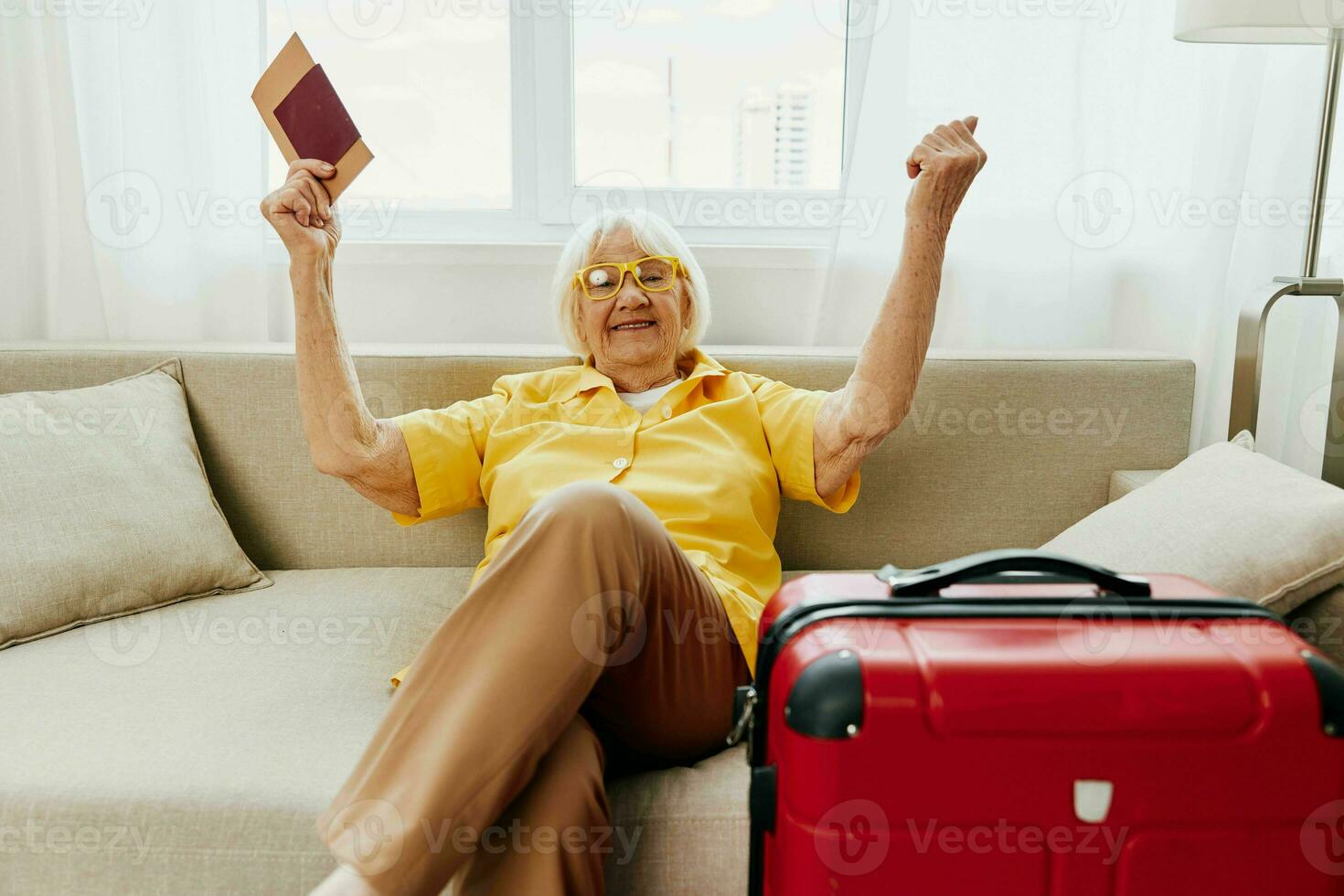 Happy senior woman with passport and travel ticket packed a red suitcase, vacation and health care. Smiling old woman joyfully sitting on the sofa before the trip raised her hands up in joy. photo