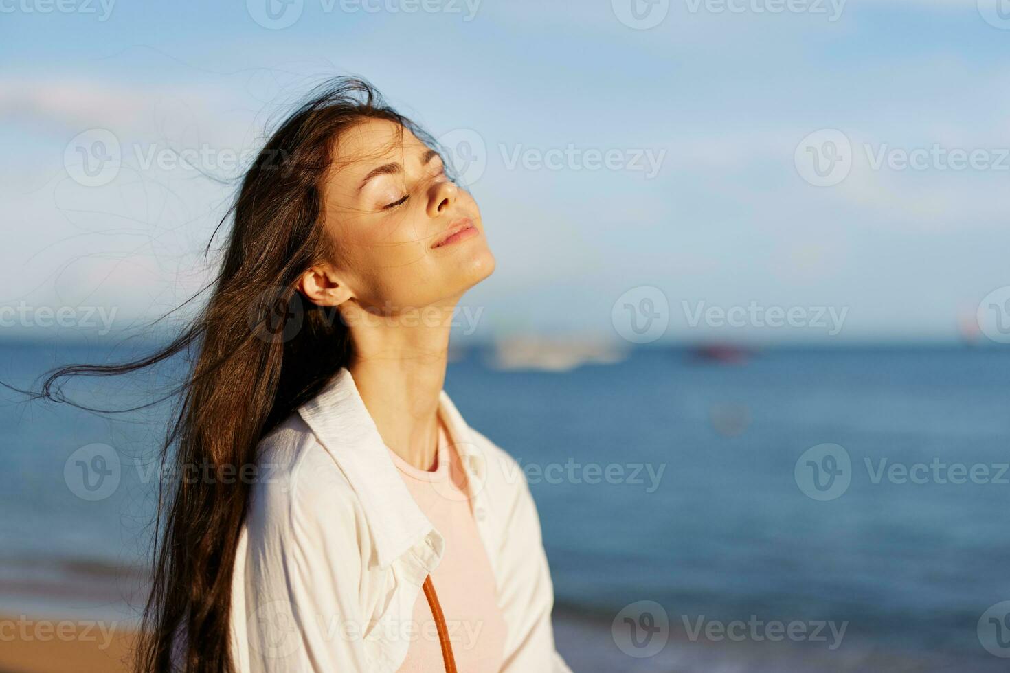 A girl with her eyes closed in the sun against the ocean smile with teeth, flying hair, tanned skin, rest, the concept of skin care in summer and spring. photo