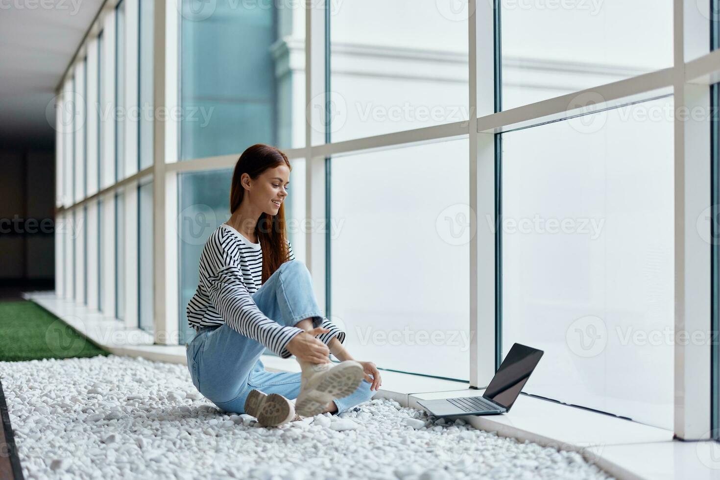 A woman smiling with teeth sits with a laptop by the big windows of a building in the city, the concept of work and leisure in the big city photo