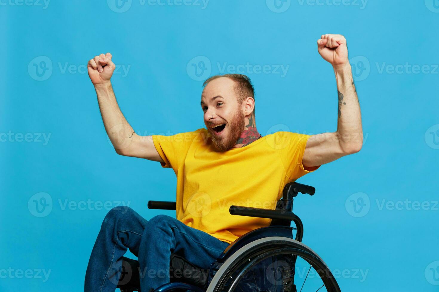 A man in a wheelchair happiness raised his hands up in a t-shirt with tattoos on his hands sits on a blue studio background, a full life, a real person photo