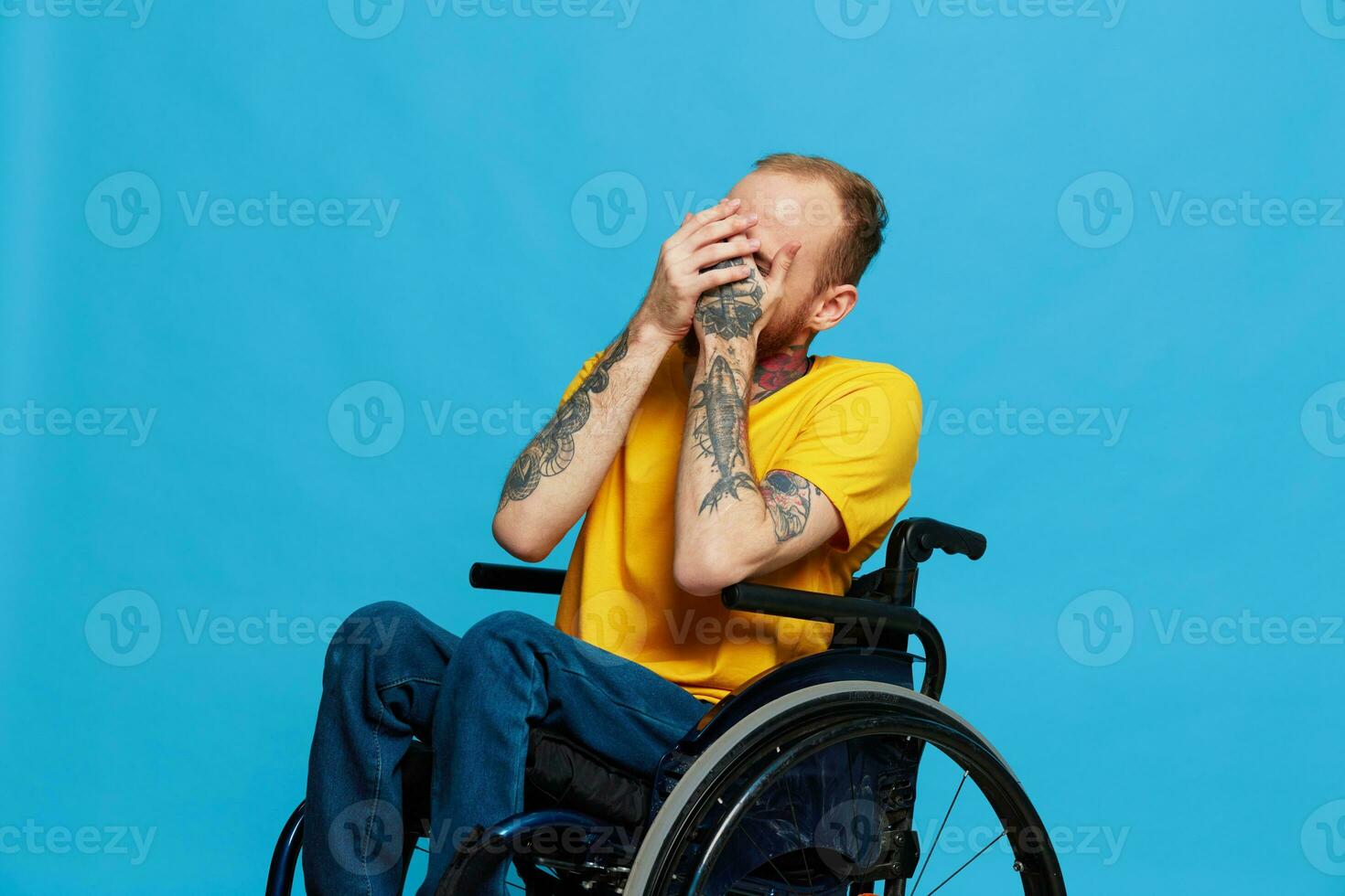 A man in a wheelchair smile looks at the camera in a t-shirt with tattoos on his arms sits on a blue studio background, a full life, a real person photo