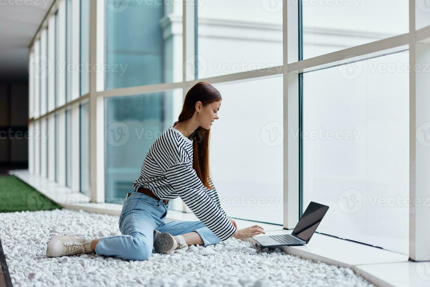 A woman sits in an office lobby by large windows and works on her laptop online via the Internet, freelance work anywhere in the world photo