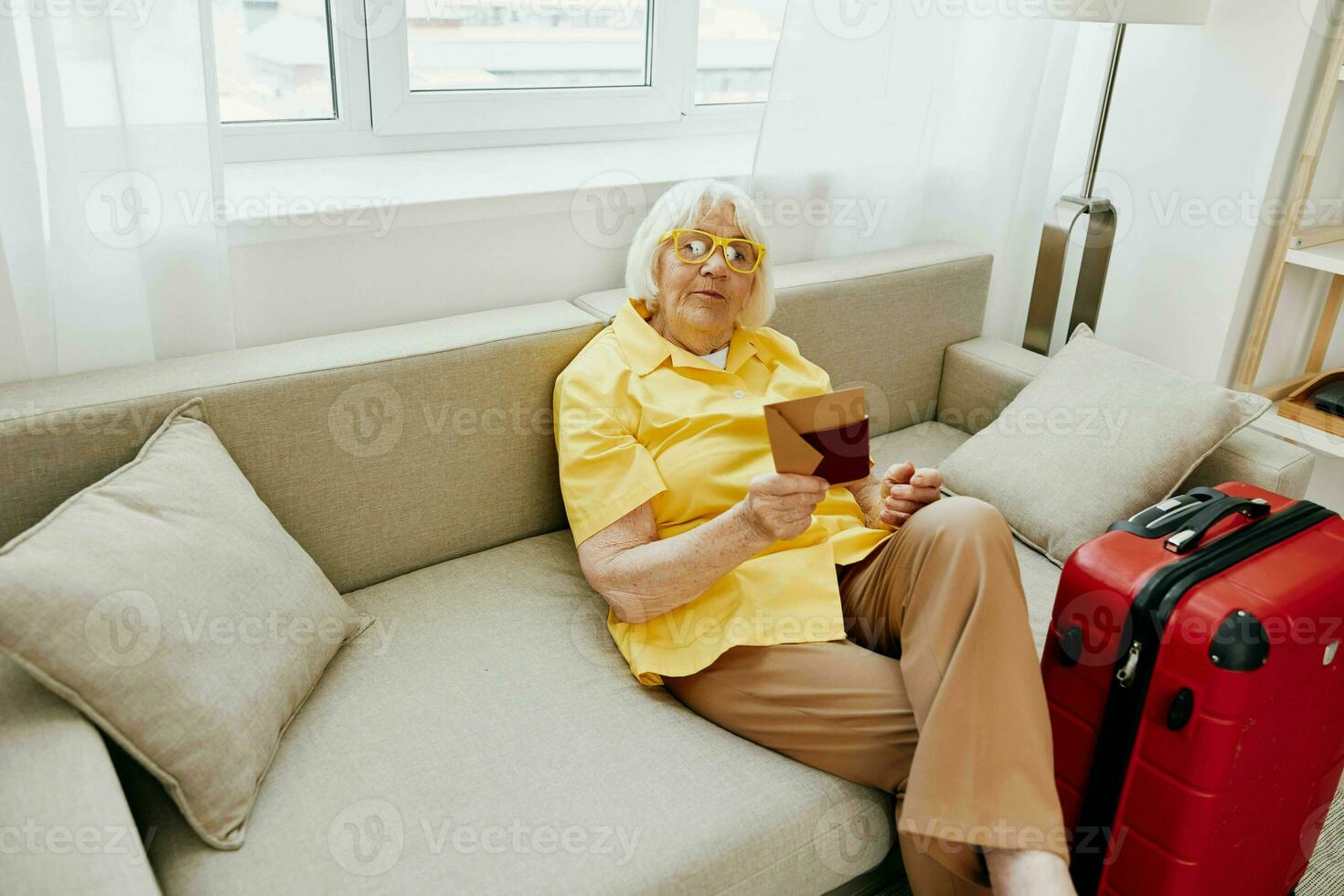 Happy senior woman with passport and travel ticket packed a red suitcase, vacation and health care. Smiling old woman joyfully sitting on the sofa before the trip raised her hands up in joy. photo