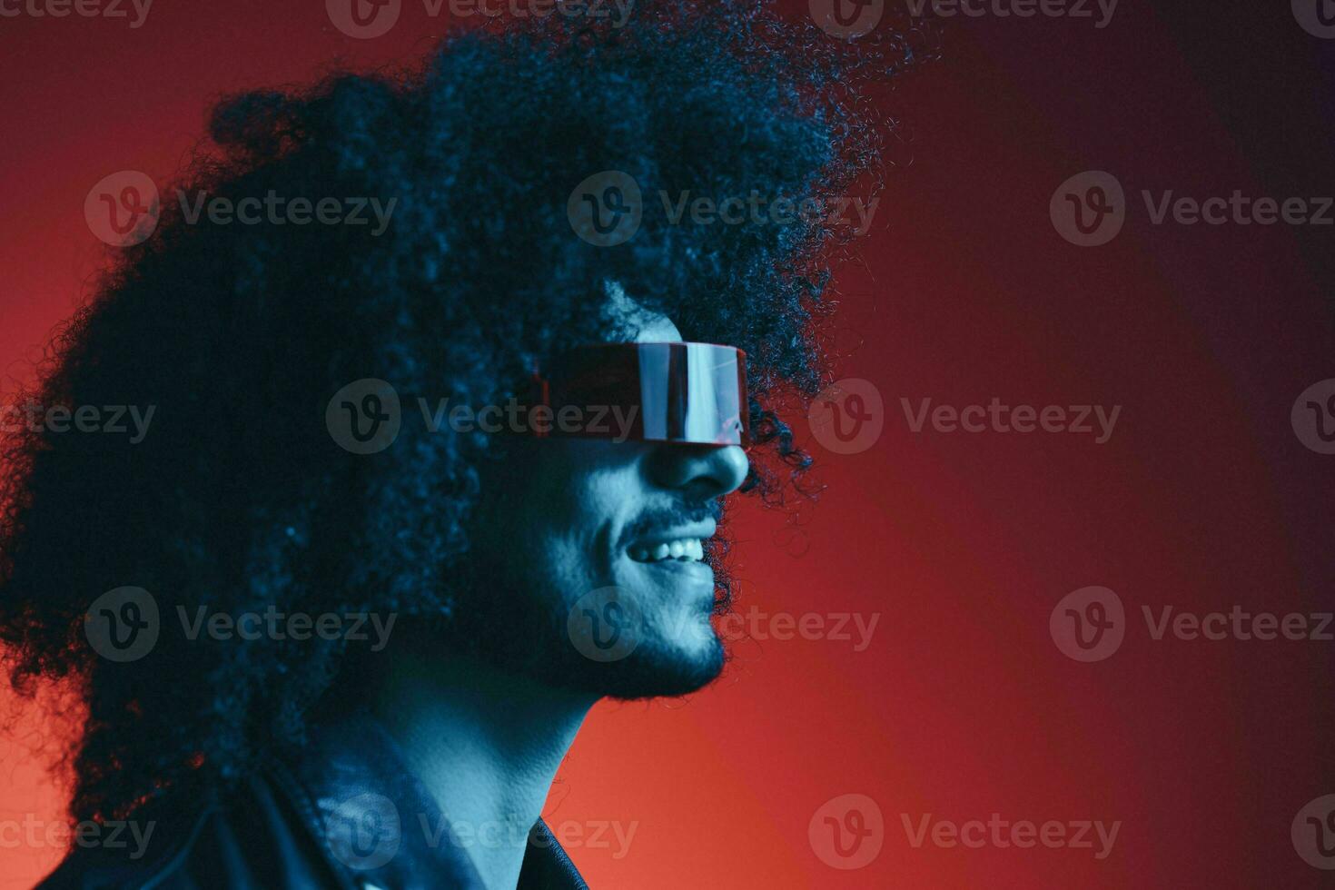 Portrait of fashion man with curly hair on red background with stylish glasses, multicultural, colored light, black leather jacket trend, modern concept. photo