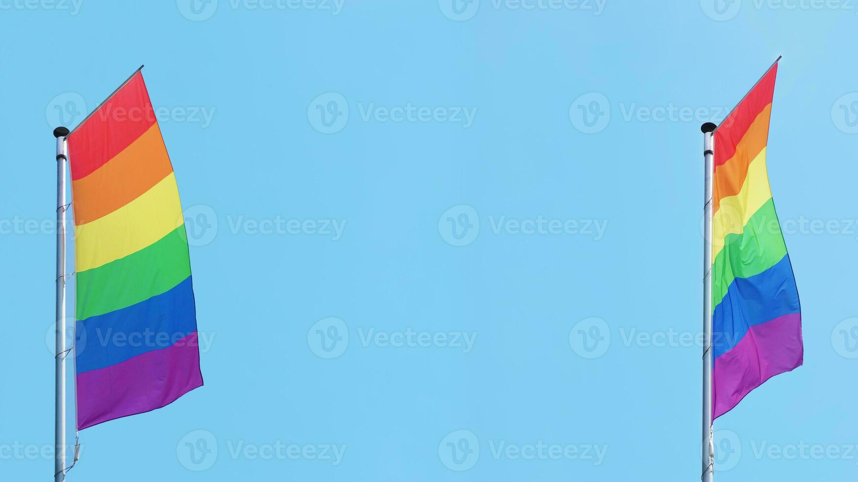 two vertical rainbow or lgbtq pride flags fly on flagpole against blue sky with copy space photo