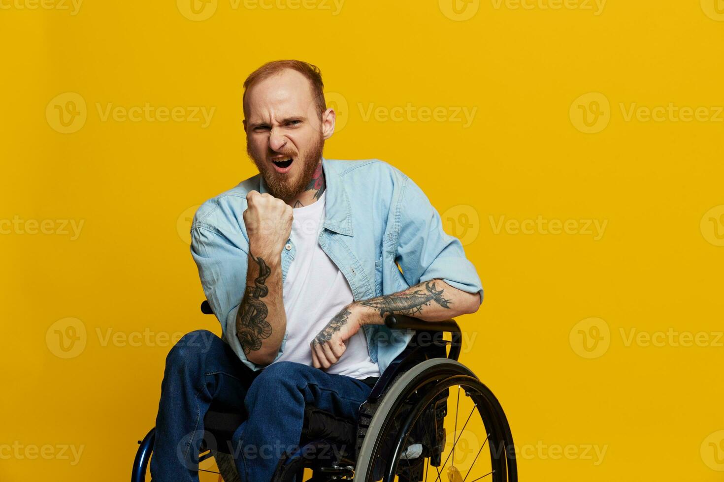 man in a wheelchair screams angrily, shows fist looks at the camera is not pleased, with tattoos on his hands sits on yellow background of the studio, the concept of health is person with disabilities photo