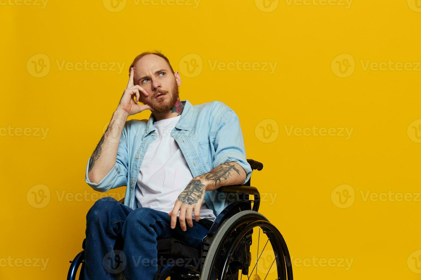 A man in a wheelchair problems with the musculoskeletal system looks at the camera thoughtfully with tattoos on his arms sits on a yellow studio background, health concept a person with disabilities photo
