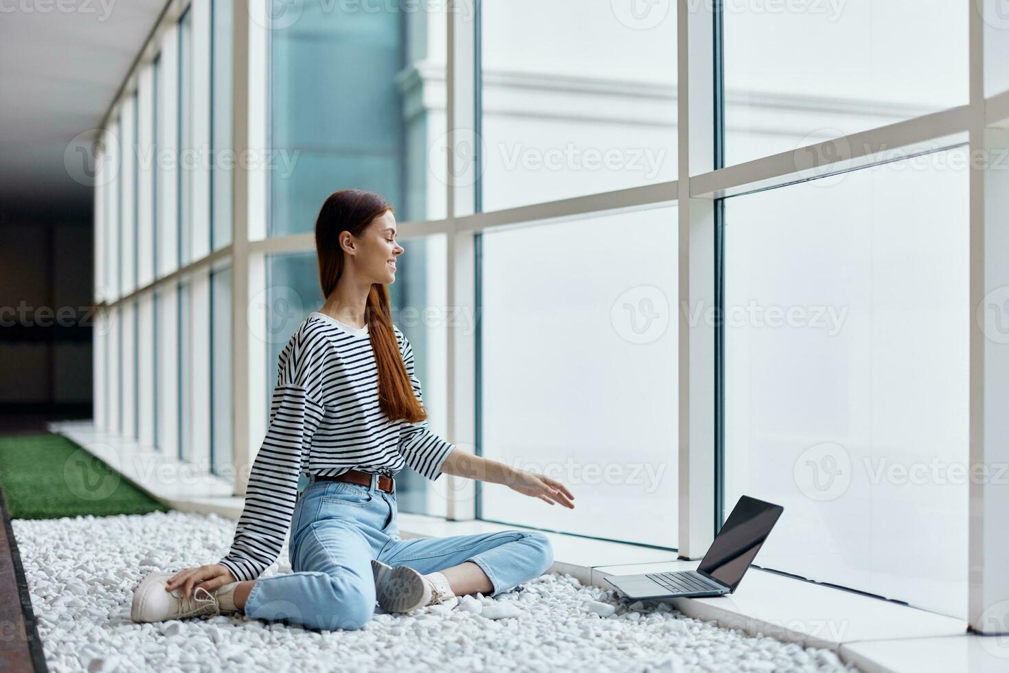 A woman smiling with teeth sits with a laptop by the big windows of a building in the city, the concept of work and leisure in the big city photo