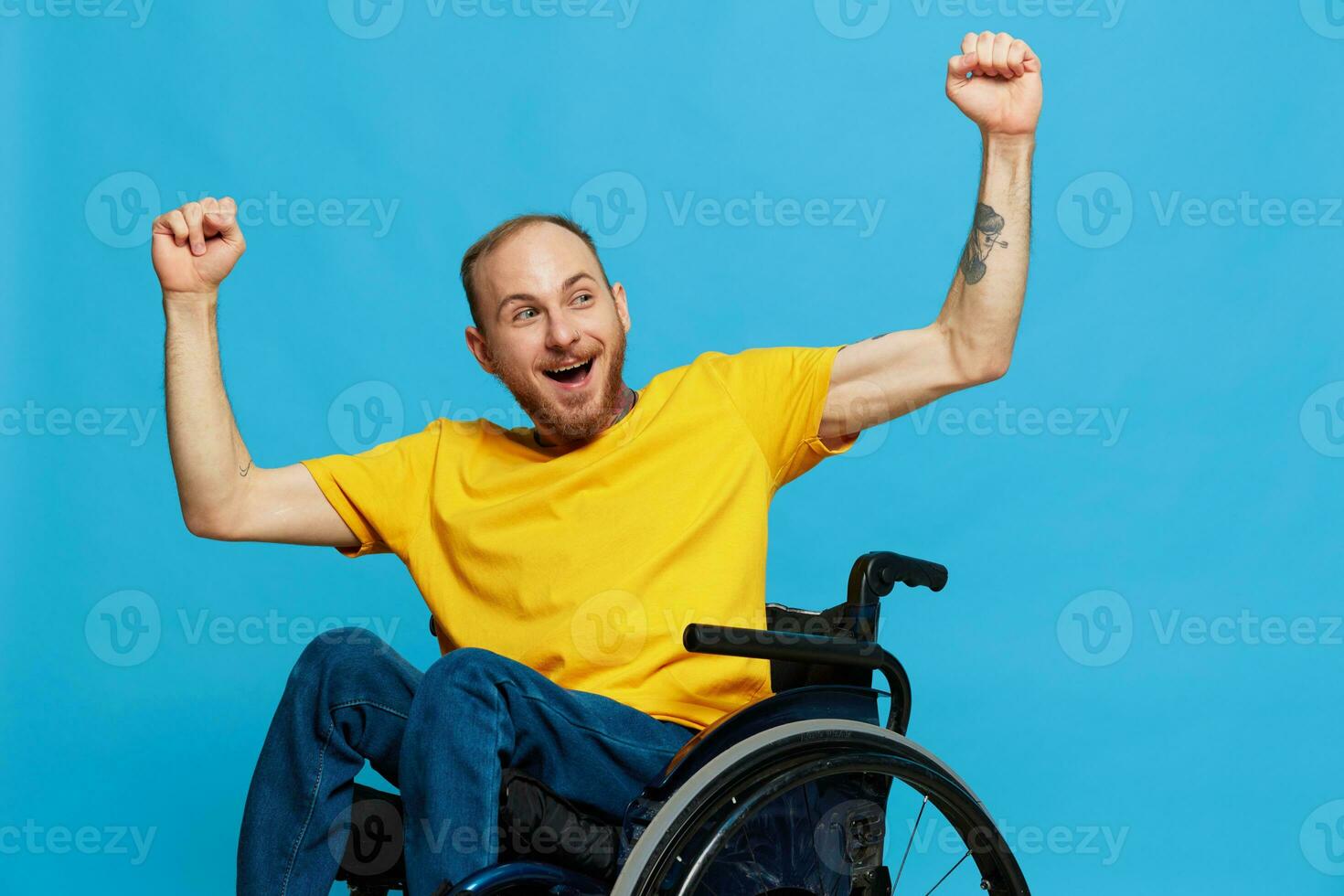 A man in a wheelchair happiness raised his hands up in a t-shirt with tattoos on his hands sits on a blue studio background, a full life, a real person photo