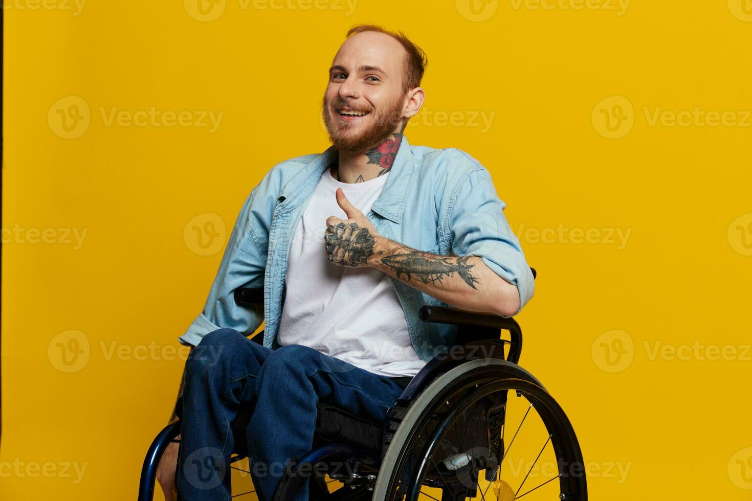A man in a wheelchair smile and happiness, thumb up, with tattoos on his hands sits on a yellow studio background, the concept of health a person with disabilities photo