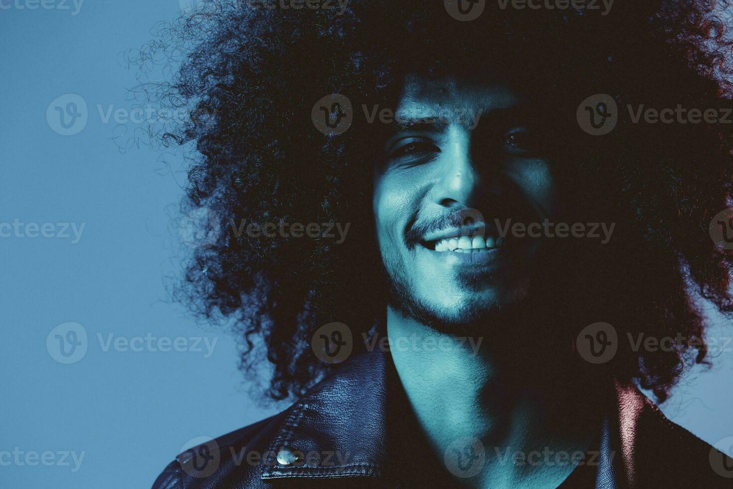 Portrait of fashion man with curly hair on blue background multinational, colored light, black leather jacket trend, modern concept. photo
