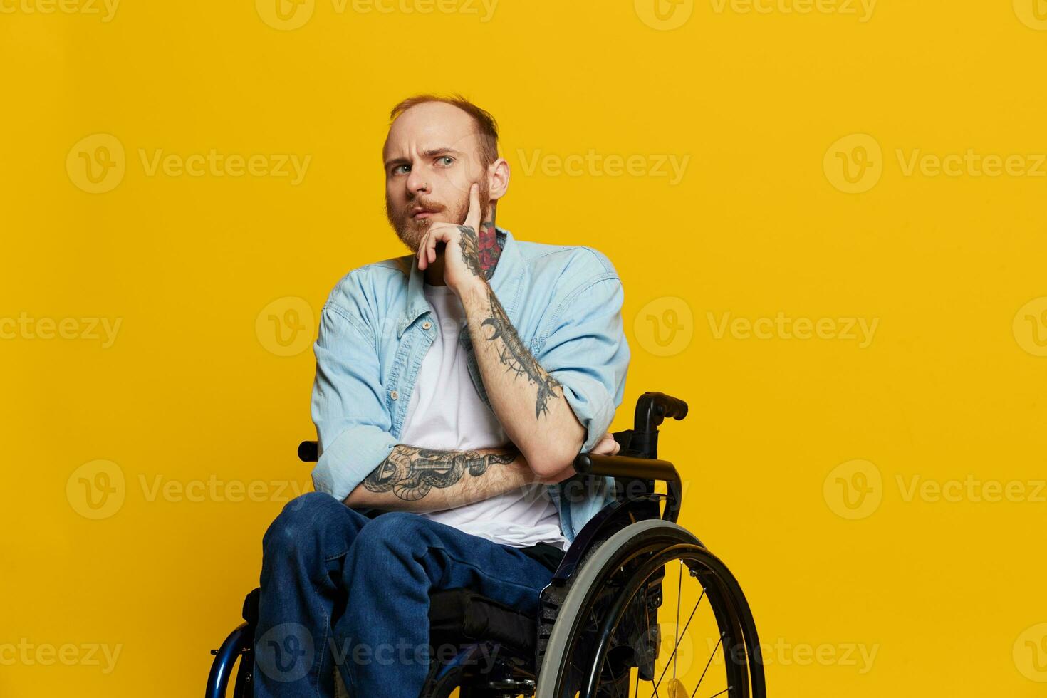 A man in a wheelchair problems with the musculoskeletal system looks at the camera shows a finger on, with tattoos on his hands sits on a yellow studio background, health concept man with disabilities photo