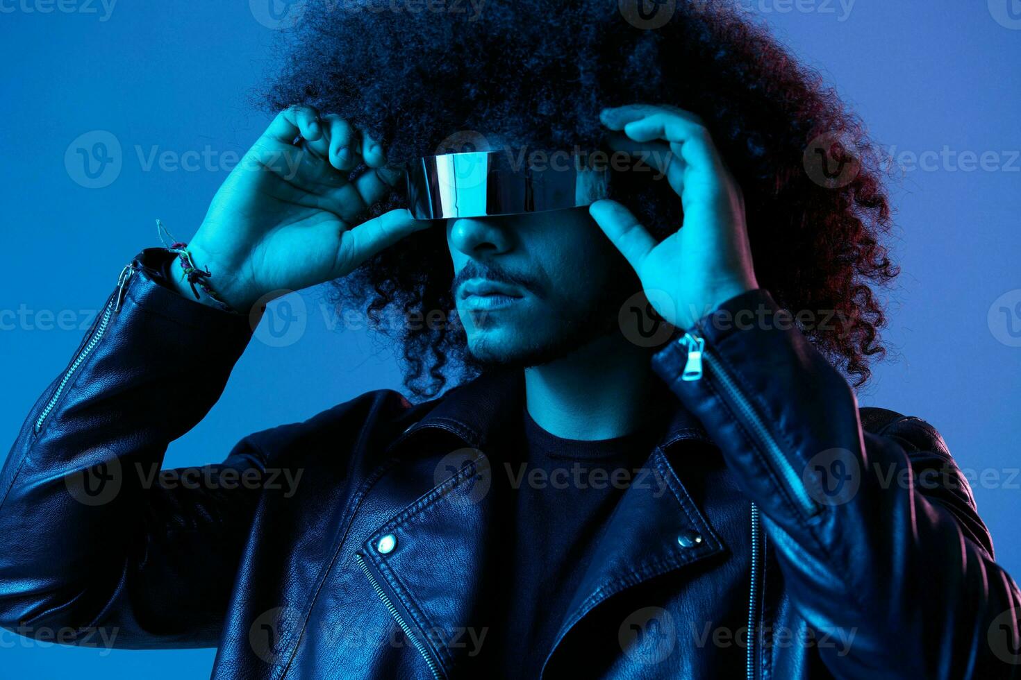 Portrait of fashion man with curly hair with stylish glasses on blue background multinational, colored light, black leather jacket trend, modern concept. photo