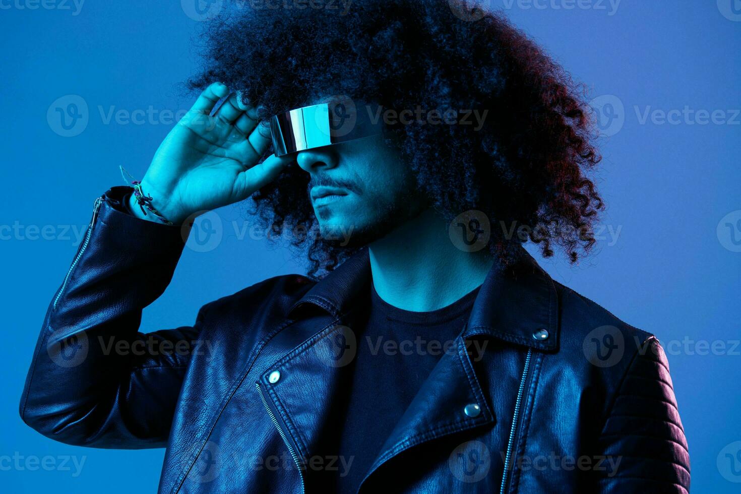 Portrait of fashion man with curly hair with stylish glasses on blue background multinational, colored light, black leather jacket trend, modern concept. photo