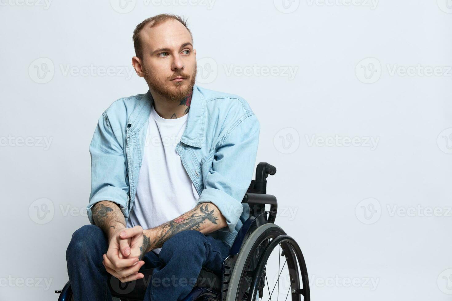 A man in a wheelchair looks thoughtfully at the camera, copy space, with tattoos on his arms sits on a gray studio background, the concept of health a person with disabilities, a real person photo