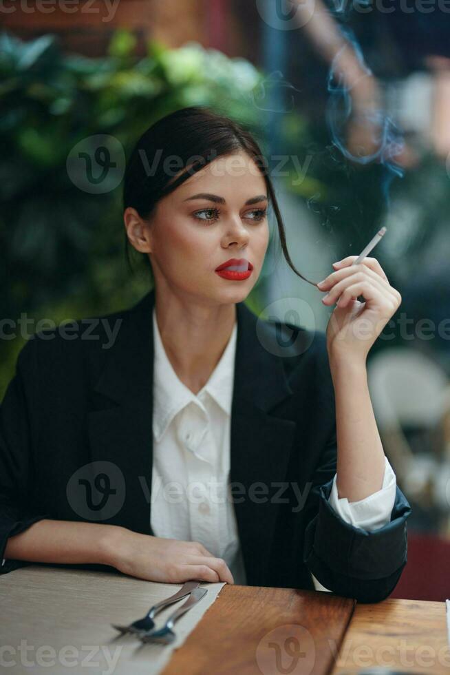 Stylish fashion woman portrait sitting in a cafe at a table and smoking a cigarette releasing smoke from her mouth with red lipstick, bad habit, smile with teeth and pensive look, cinematic vintage photo