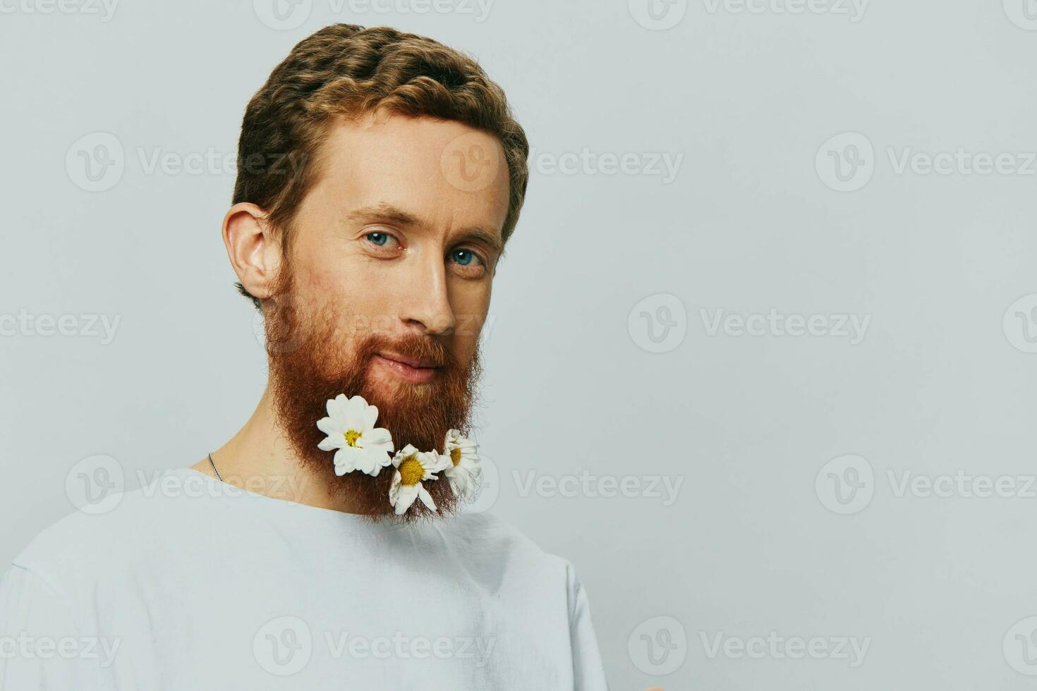 Portrait of a funny man in a white T-shirt with flowers daisies in his beard on a white isolated background, copy place. Holiday concept and congratulations. photo
