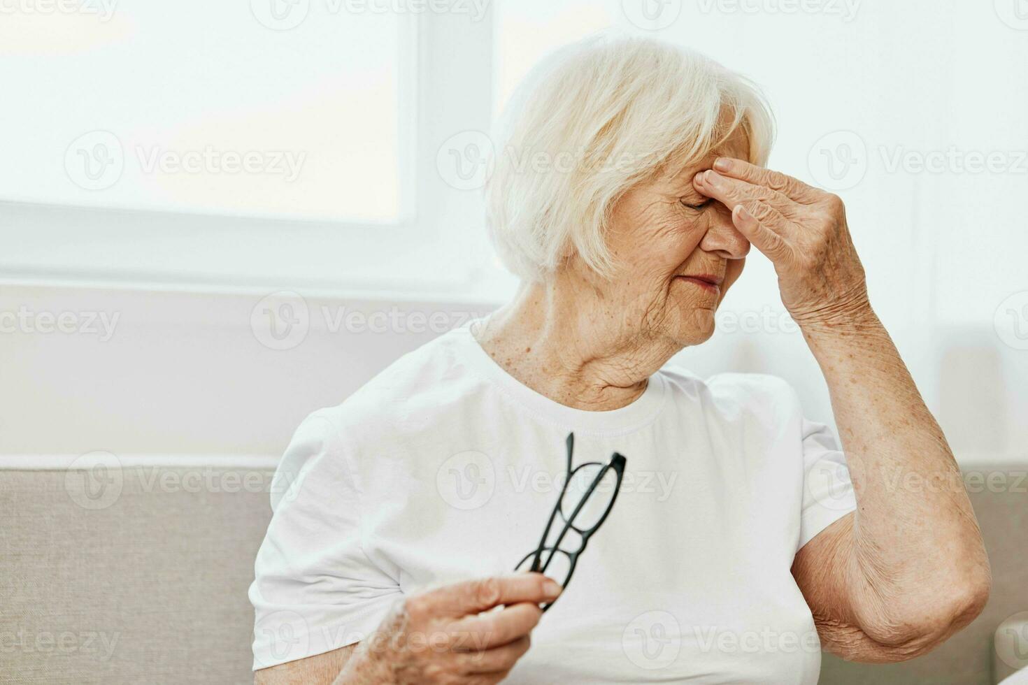 Elderly woman severe eye pain sitting on the couch, health problems in old age, poor quality of life. A grandmother with gray hair holds on to her sore eyes with glasses in her hand. photo