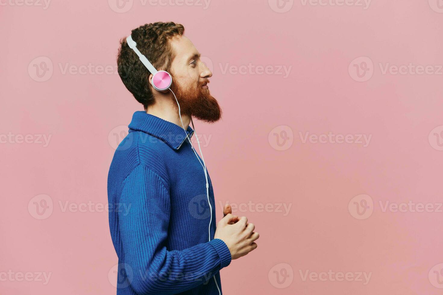 Portrait of a redheaded man wearing headphones smiling and dancing, listening to music on a pink background. A hipster with a beard. photo